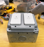 A gray dual-gang electrical junction box with two white rocker switches mounted on top, set on a wooden workbench with tools and electronic components in the background.